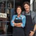 Two cheerful small business owners smiling and looking at camera while standing at entrance door. Happy mature man and mid woman at entrance of newly opened restaurant with open sign board.